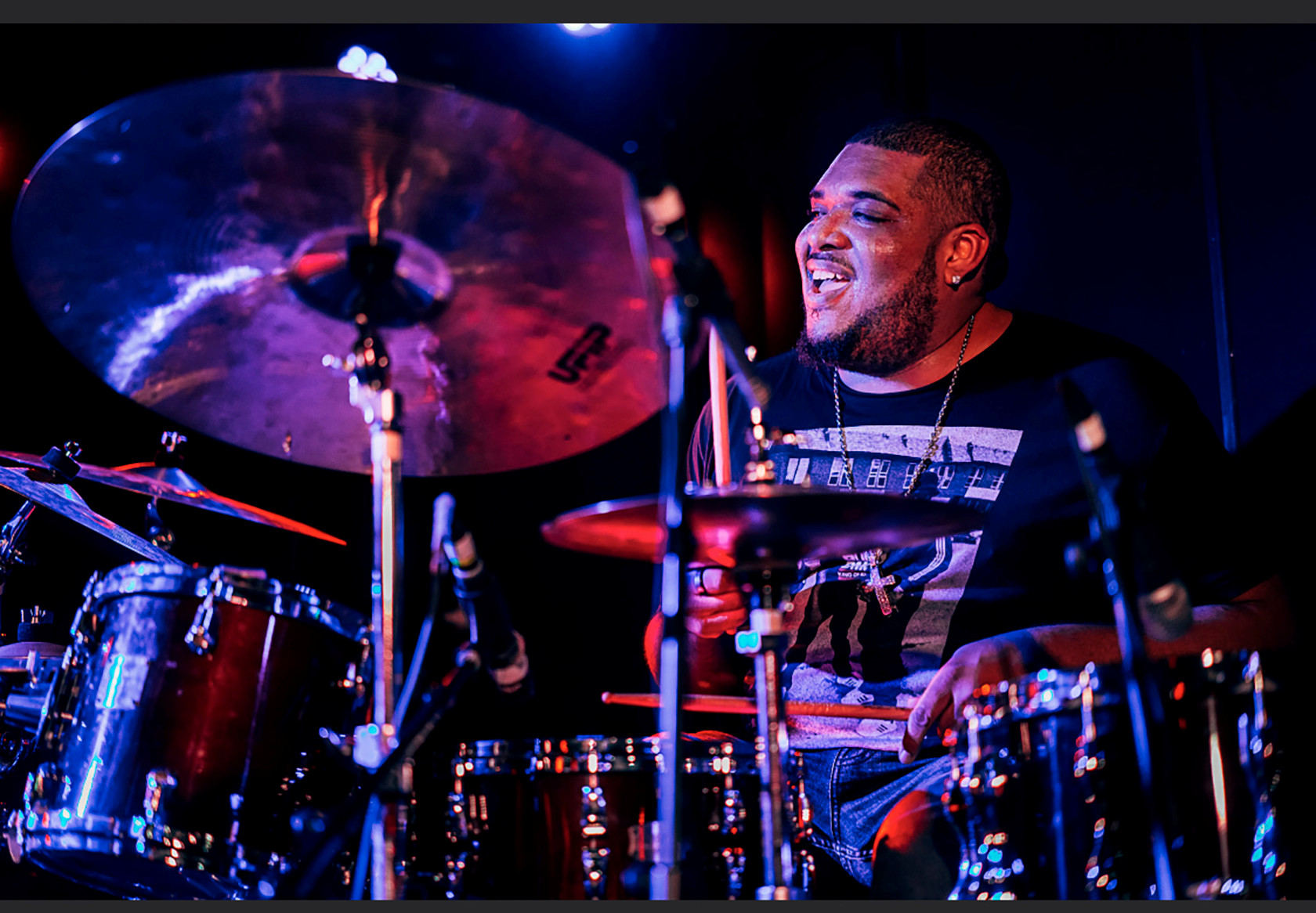 a man plays the drums on stage happily with dramatic red and blue lighting 