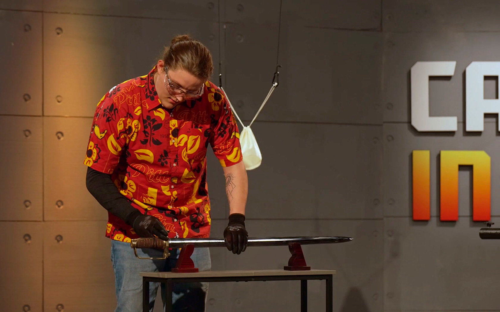 a male student wearing safety goggles is looking down at a sword laying on a stand on a table