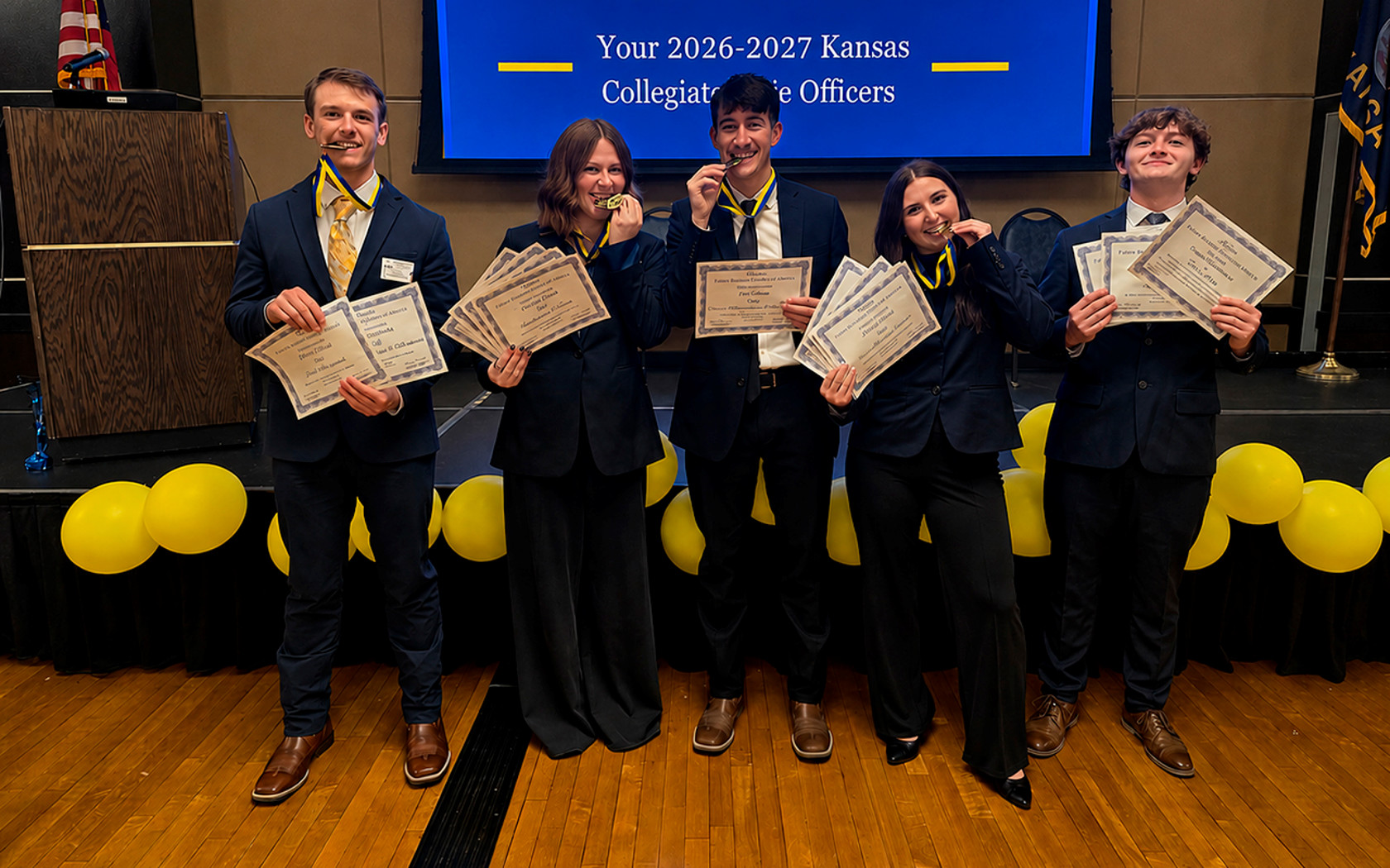 a group of students dressed in suits holding certificates and wearing medals, which they are biting between their teeth as they smile