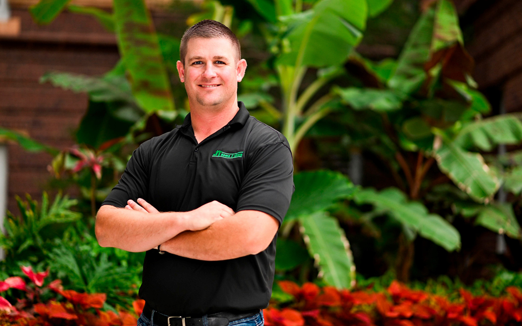 man wearing black shirt with company logo, crossing arms, standing in front of tropical plants, smiling
