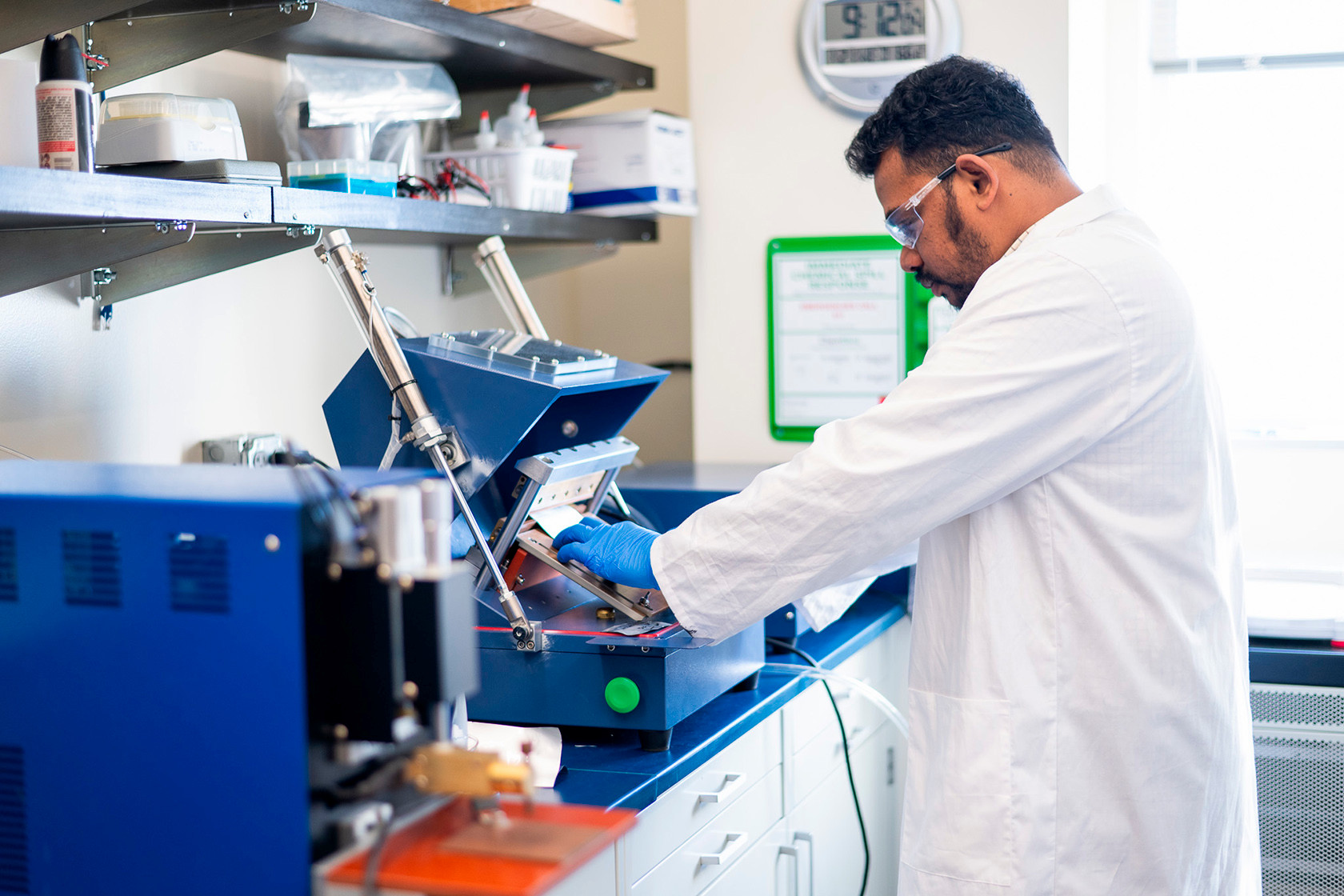 A man looks at a scientific machine in white coat and goggles